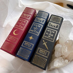 Puede incluir: Tres libros de tapa dura con letras doradas e ilustraciones. Los libros se titulan "House of Earth and Blood", "House of Sky and Breath" y "House of Flame and Shadow". Los tres libros forman parte de la serie Crescent City de Sarah J. Maas.