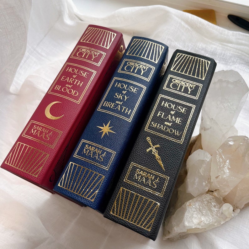 Puede incluir: Tres libros de tapa dura con letras doradas e ilustraciones. Los libros se titulan "House of Earth and Blood", "House of Sky and Breath" y "House of Flame and Shadow". Los tres libros forman parte de la serie Crescent City de Sarah J. Maas.