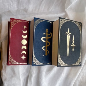 Puede incluir: Tres libros de tapa dura con detalles dorados y dise&ntilde;os decorativos. El libro de la izquierda tiene una cubierta roja con medias lunas y estrellas doradas. El libro del medio tiene una cubierta azul con un dise&ntilde;o de serpiente dorada. El libro de la derecha tiene una cubierta azul con un dise&ntilde;o de espada dorada.