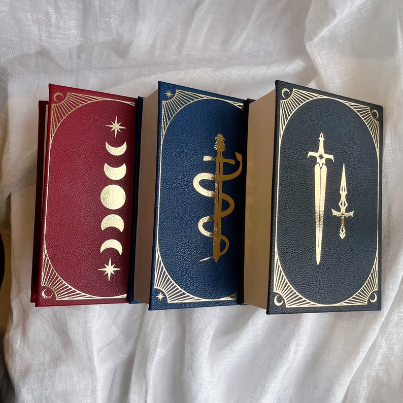 Puede incluir: Tres libros de tapa dura con detalles dorados y dise&ntilde;os decorativos. El libro de la izquierda tiene una cubierta roja con medias lunas y estrellas doradas. El libro del medio tiene una cubierta azul con un dise&ntilde;o de serpiente dorada. El libro de la derecha tiene una cubierta azul con un dise&ntilde;o de espada dorada.