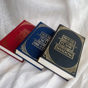 Puede incluir: Tres libros de tapa dura con letras doradas en el lomo. Los libros se titulan "House of Earth and Blood", "House of Sky and Breath" y "House of Flame and Shadow".