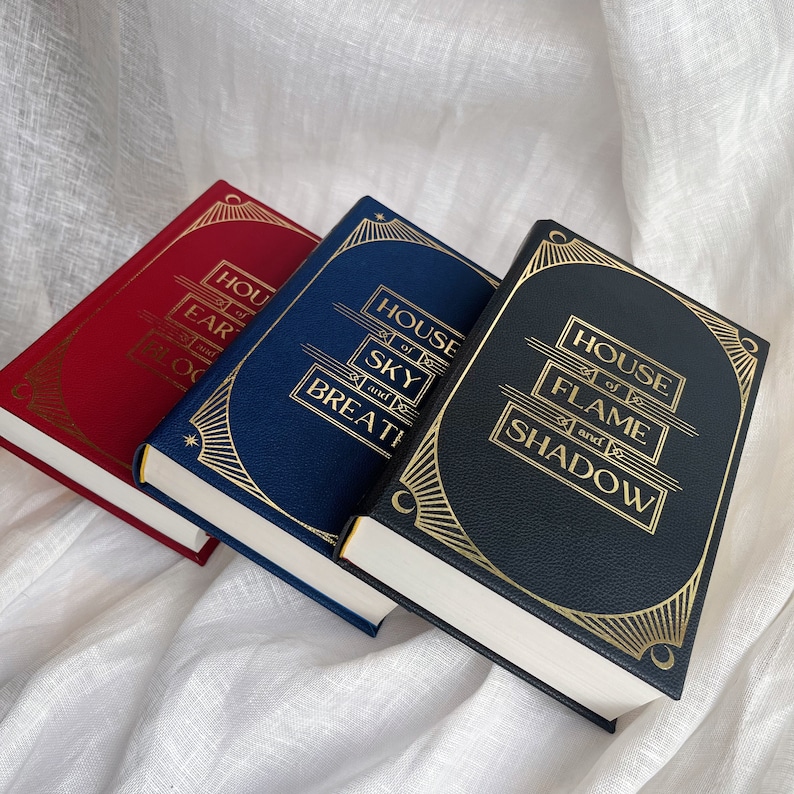 Puede incluir: Tres libros de tapa dura con letras doradas en el lomo. Los libros se titulan "House of Earth and Blood", "House of Sky and Breath" y "House of Flame and Shadow".