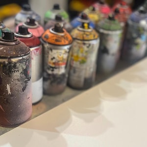 May include: A selection of used spray paint cans in diverse colours, such as blue, brown, pink, and orange. The cans are arranged in a row, with a white surface in the foreground. The cans show signs of use, with paint splatters and wear.