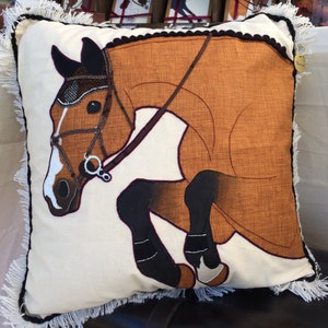 May include: A brown and white pillow with a brown horse jumping over a fence. The horse is wearing a bridle and a black nose net. The pillow has a white fringe around the edges.