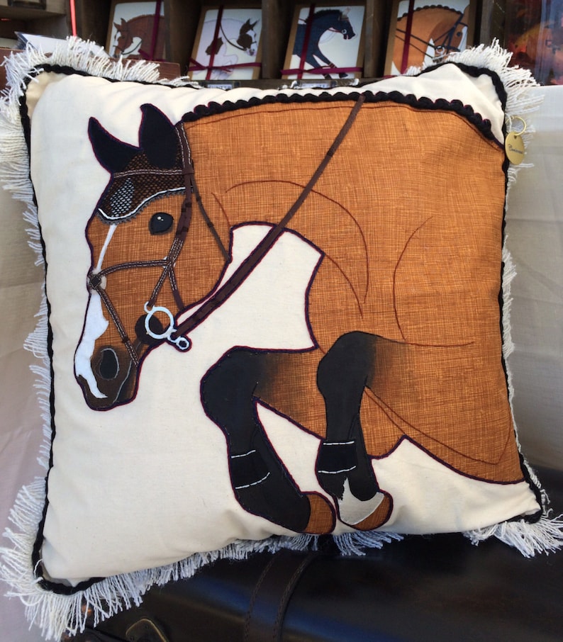 May include: A brown and white pillow with a brown horse jumping over a fence. The horse is wearing a bridle and a black nose net. The pillow has a white fringe around the edges.