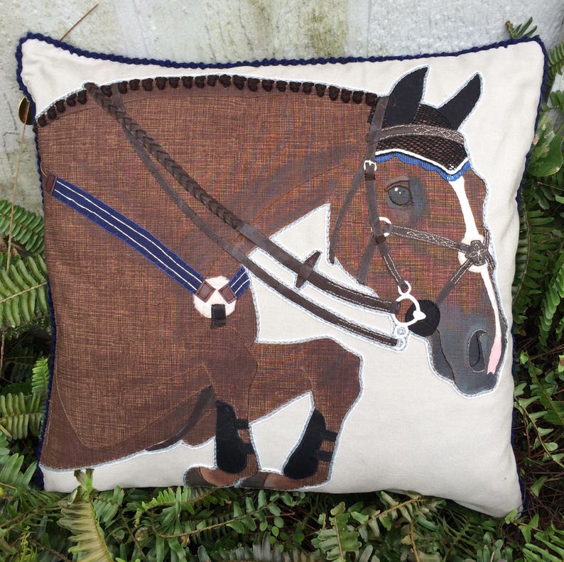 May include: A brown horse head with a black bridle and a blue and white browband is stitched onto a white pillow with a brown and white patterned fabric. The pillow has a navy blue trim.