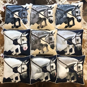 May include: Nine decorative throw pillows featuring a white background and a horse jumping over a fence. The horses are all different colours, including grey, white, and black. The pillows have a white trim and a blue background.