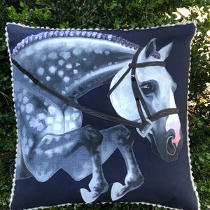 May include: A navy blue cushion with a white horse jumping over a fence. The horse has a black bridle and a white and grey spotted coat. The cushion has a white trim.