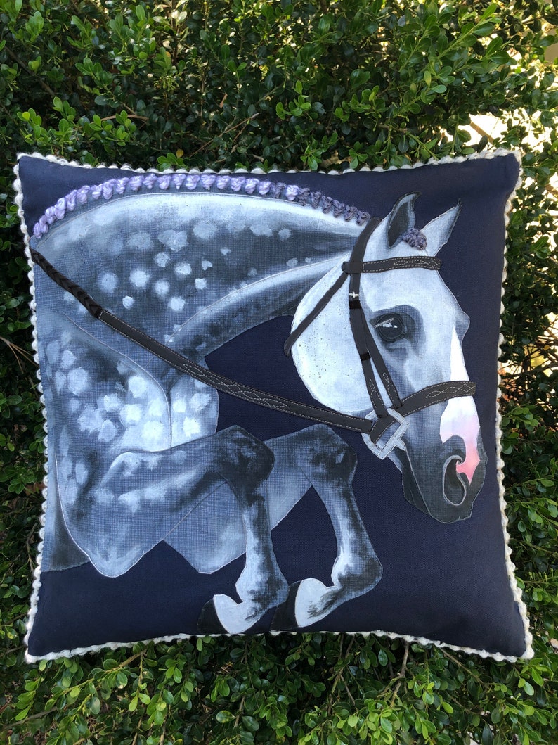 May include: A navy blue cushion with a white horse jumping over a fence. The horse has a black bridle and a white and grey spotted coat. The cushion has a white trim.
