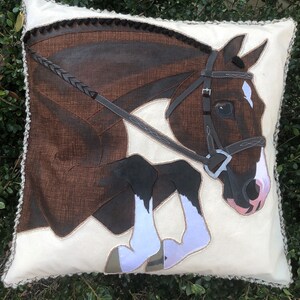 May include: A white cushion featuring a brown and white horse leaping over a brown leather bridle. The horse has a white star on its forehead and white socks on its legs.