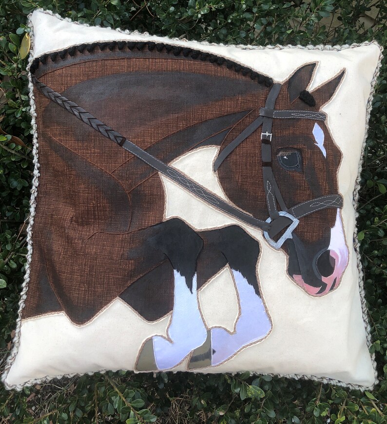 May include: A white cushion featuring a brown and white horse leaping over a brown leather bridle. The horse has a white star on its forehead and white socks on its legs.