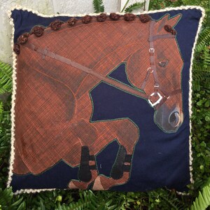 May include: A brown horse with a black bridle and black boots is depicted on a navy blue pillow with a white crocheted border.