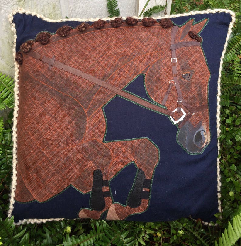 May include: A brown horse with a black bridle and black boots is depicted on a navy blue pillow with a white crocheted border.