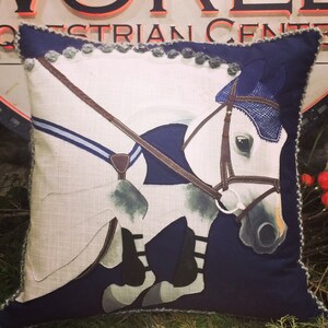 May include: A decorative pillow with a white horse jumping over a blue and brown fence. The pillow has a navy blue background and a gray and blue trim.