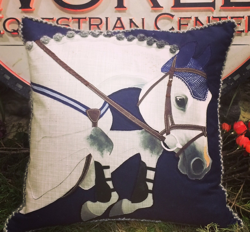 May include: A decorative pillow with a white horse jumping over a blue and brown fence. The pillow has a navy blue background and a gray and blue trim.