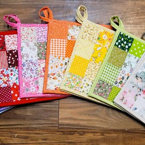 May include: A collection of seven square fabric pot holders arranged in a fan shape. Each pot holder features a unique patchwork design with various floral, gingham, and polka dot patterns in blue, purple, red, pink, orange, yellow, green, and white.