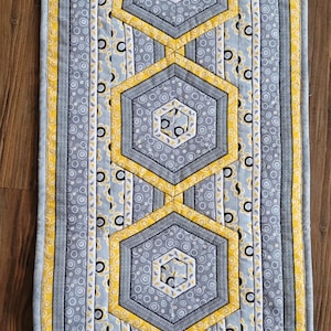 May include: A gray and yellow table runner with a geometric pattern of hexagons. The runner has a scalloped edge and is made of fabric.