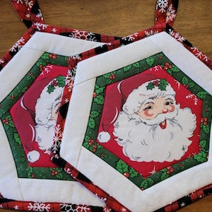 May include: Two Christmas-themed pot holders. Each has a red center with a Santa Claus face, surrounded by green holly and white borders. The edges are red and black plaid with snowflake accents.