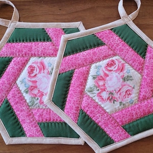 May include: Two quilted, hexagonal pot holders. Each features a central floral design with pink roses, surrounded by pink and green fabric sections. Beige binding and a loop for hanging complete the design.