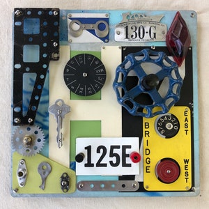 May include: A colorful metal wall art piece with various industrial and mechanical elements, including a blue valve, a dial, a key, and a number plate with the text "125E".
