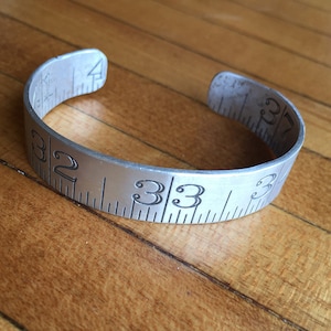 May include: A silver-colored cuff bracelet with engraved ruler markings and numbers. The bracelet has a vintage, industrial aesthetic, with measurements from 32 to 34 inches. The bracelet is open at the ends.