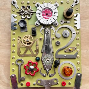 May include: A unique assemblage art piece on a light green background. It features various metal objects, including gears, keys, and a "WALK-A-MATIC" dial. The composition includes a red flower-shaped valve and a number "2" made of metal.