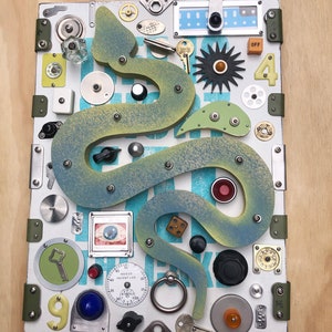 May include: A wooden board with a blue and green snake design. The board is covered in various objects, including keys, knobs, buttons, gears, and a compass. The board is a tactile learning tool for children.