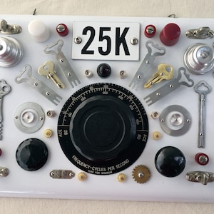 May include: A white metal panel with various metal and plastic objects attached, including keys, gears, knobs, and a dial labeled "FREQUENCY-CYCLES PER SECOND". The panel also has a white label with the text "25K" and a smaller white label with the text "319".