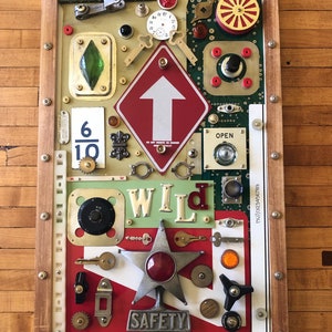 May include: A framed mixed media artwork featuring a variety of vintage and industrial objects, including a red traffic sign with a white arrow pointing upwards, a star-shaped object with a red light, and the word "WILD" in white letters on a green background. The artwork also includes a variety of other objects, such as keys, gears, and a clock face.