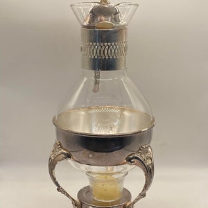 May include: Silver-plated glass tea pot with a glass base and a candle warmer. The tea pot has a decorative design on the top and a handle on the side.