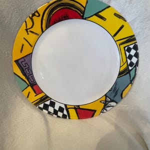 May include: A decorative ceramic plate with a white center and a vibrant yellow border. The border features a colorful geometric design with red, black, teal, and white shapes. The plate is round and suitable for serving food.