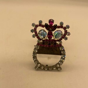 May include: A silver-toned brooch with a crown design. The crown is adorned with pink and clear rhinestones. The brooch has a circular mirror in the center.