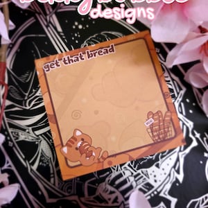 May include: A square sticky note with a brown and orange color scheme featuring the text get that bread at the top. The design includes a cartoon red panda eating a loaf of bread and a small basket of bread loaves in the bottom right corner.