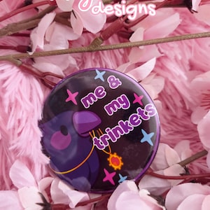 May include: A round button with the text "me & my trinkets" and a cartoon bird illustration. The button is primarily black with purple, pink, and blue accents. The background features pink flowers and a pink, fluffy texture.