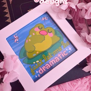 May include: A framed art print featuring a cartoon frog on a lily pad, with the text "just let me be dramatic". The frog is green and yellow with a pink mouth, set against a blue background. The frame is white, and the artwork is surrounded by pink flowers.
