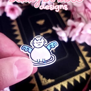 May include: A holographic sticker featuring a white cat with blue wings and a halo above its head. The sticker has a white outline and displays the text bekkybubble designs in a pink font at the top of the image, set against a dark background.