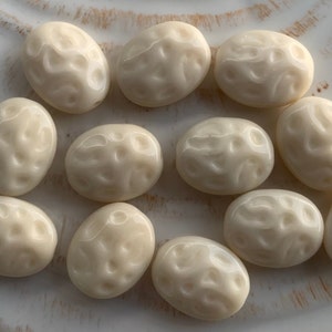 May include: A collection of off-white oval-shaped beads with a textured surface. The beads have a glossy finish and are arranged on a white surface.