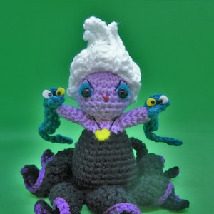 Puede incluir: Una muñeca Ursula de ganchillo de La Sirenita. Tiene la piel morada, el pelo blanco y tentáculos negros y morados. Está sosteniendo dos anguilas verdes y azules en sus manos.