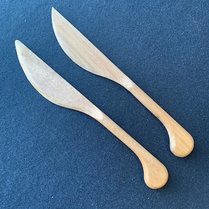 Può includere: Due coltelli da burro in legno con manici arrotondati. I coltelli sono di colore marrone chiaro e hanno una finitura liscia.