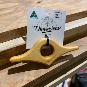 May include: A wooden book page holder made of Huon Pine, 16mm thick. The holder is shaped like a bird with a round opening in the center. The holder is sitting on a stack of wooden boards. The label reads "Timber Dimensions Est 1998" and "Huon Pine 16mm".