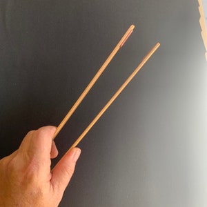 Puede incluir: Un par de palillos de madera, uno con una punta roja, sostenido en una mano sobre un fondo negro.