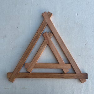 May include: A wooden sculpture with a geometric design. It features a large triangle frame with a smaller triangle inside. The sculpture is made of light brown wooden slats, creating a layered effect. The design is simple and modern.