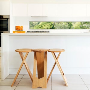 May include: Three wooden stools with round seats and angled legs. The stools are made of light-colored wood and have a minimalist design. The stools are in a modern kitchen setting.