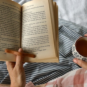 Può includere: Un libro aperto con un segnalibro in legno, una tazza di tè con un motivo floreale e una coperta a righe. Il libro è aperto su una pagina con testo. La scena suggerisce relax e lettura.