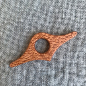 May include: A wooden bookmark with a unique, abstract shape and a hole in the center. The bookmark is made of light brown wood with a reddish-brown grain.