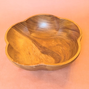 May include: A wooden bowl with a distinctive, four-lobed design. The bowl is crafted from light brown wood, showcasing a visible grain. The interior is smooth and polished, reflecting light. It rests on a peach-coloured surface.