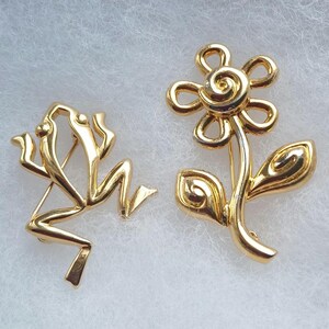 May include: Two gold-toned metal brooches. One brooch is shaped like a stylized frog with elongated limbs and a smooth, reflective surface. The second brooch is shaped like a flower with a central spiral, six rounded petals, a stem, and two leaves.