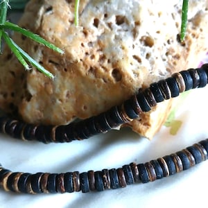May include: A necklace made of dark brown and tan wooden beads. The beads are circular and strung together to form a necklace. The necklace is resting on a light-colored surface with a rock and greenery in the background.