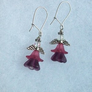 K&ouml;nnte beinhalten: Ein Paar silberne Ohrh&auml;nger mit lila Blumenanh&auml;ngern. Die Blumen sind durchscheinend und haben ein mehrschichtiges Design.
