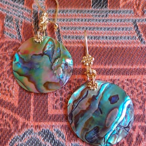 May include: A pair of gold-toned earrings with iridescent, round abalone shell pendants. The shells display a mix of blue, green, and purple hues. The earrings have a simple hook design.